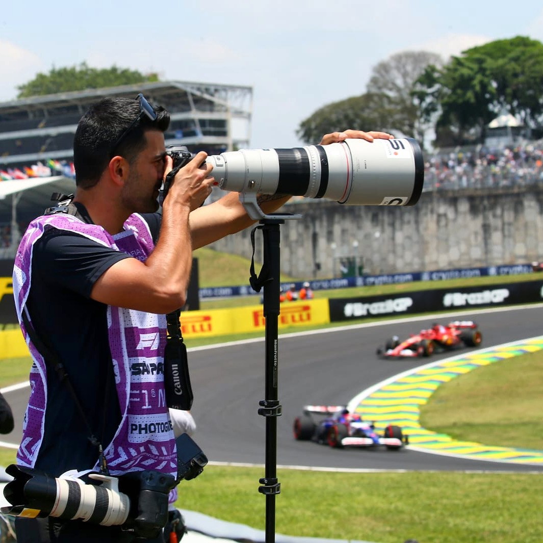 Lucas Miranda fotografando o GP de São Paulo com teleobjetiva, com carros de F1 na pista ao fundo